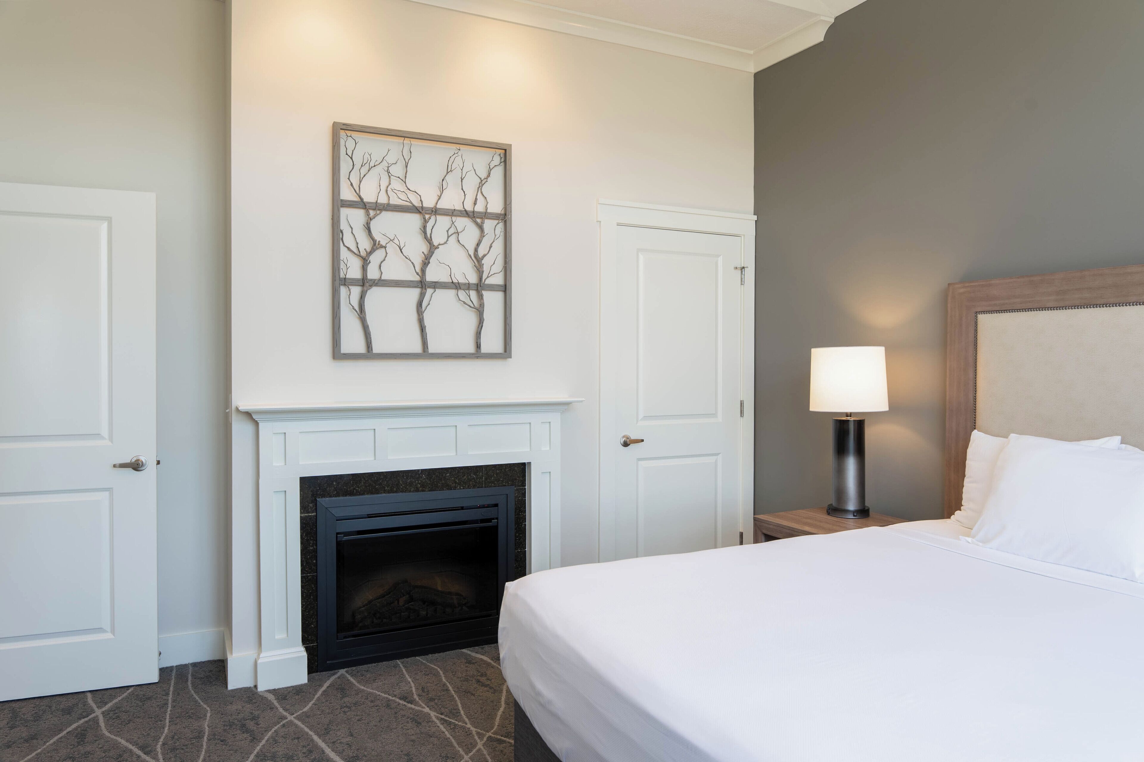 A hotel room with a fireplace, a bed with white bedding, a nightstand with a lamp, and a wall-mounted artwork.
