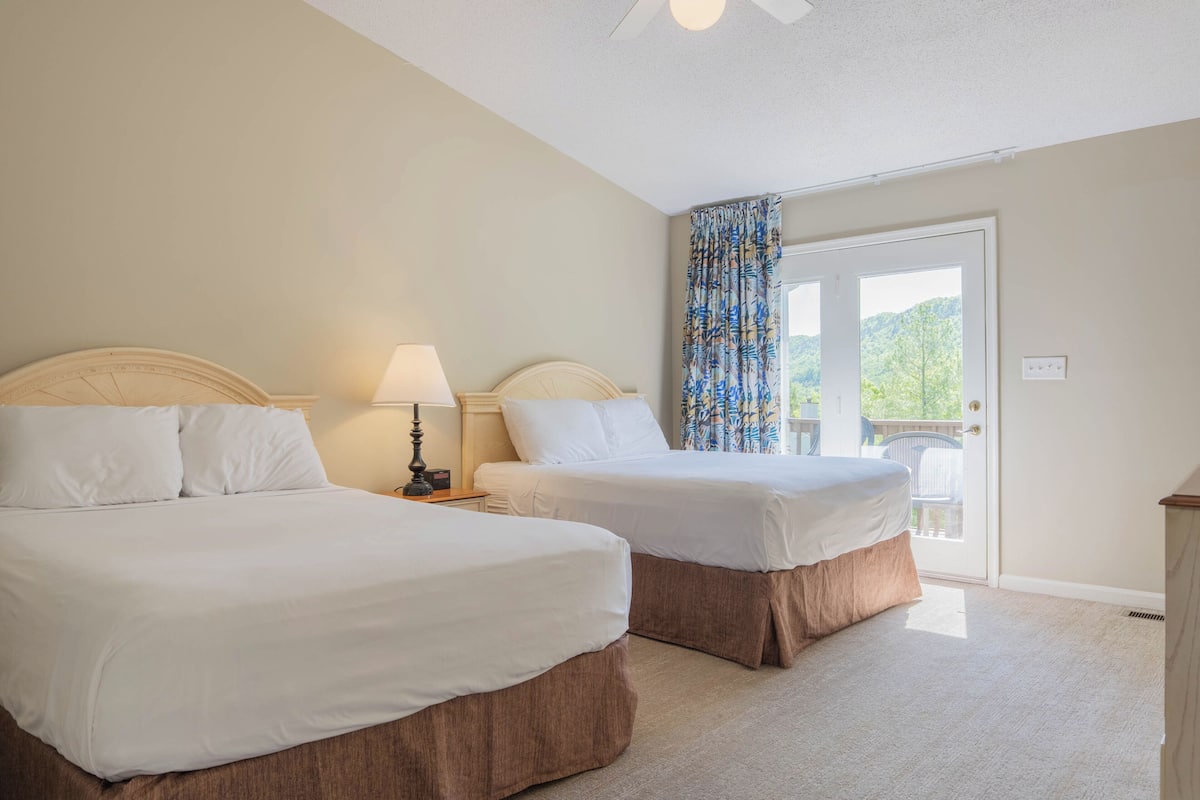 A hotel room with two beds, a ceiling fan, a nightstand with a lamp, and a view of greenery through the door.