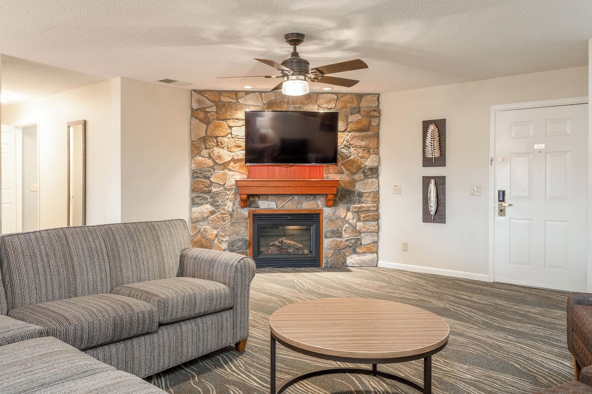 A living room with a stone fireplace, a flat-screen TV, a ceiling fan, and a patterned carpet.