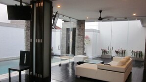 A modern living room with a white sofa, a glass coffee table, and a black chair. There is a TV mounted on the wall, and a ceiling fan is visible.