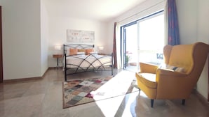 Ein Schlafzimmer mit Bett, Sessel, Nachttisch, Fenster mit Vorhängen und Deckenleuchte.