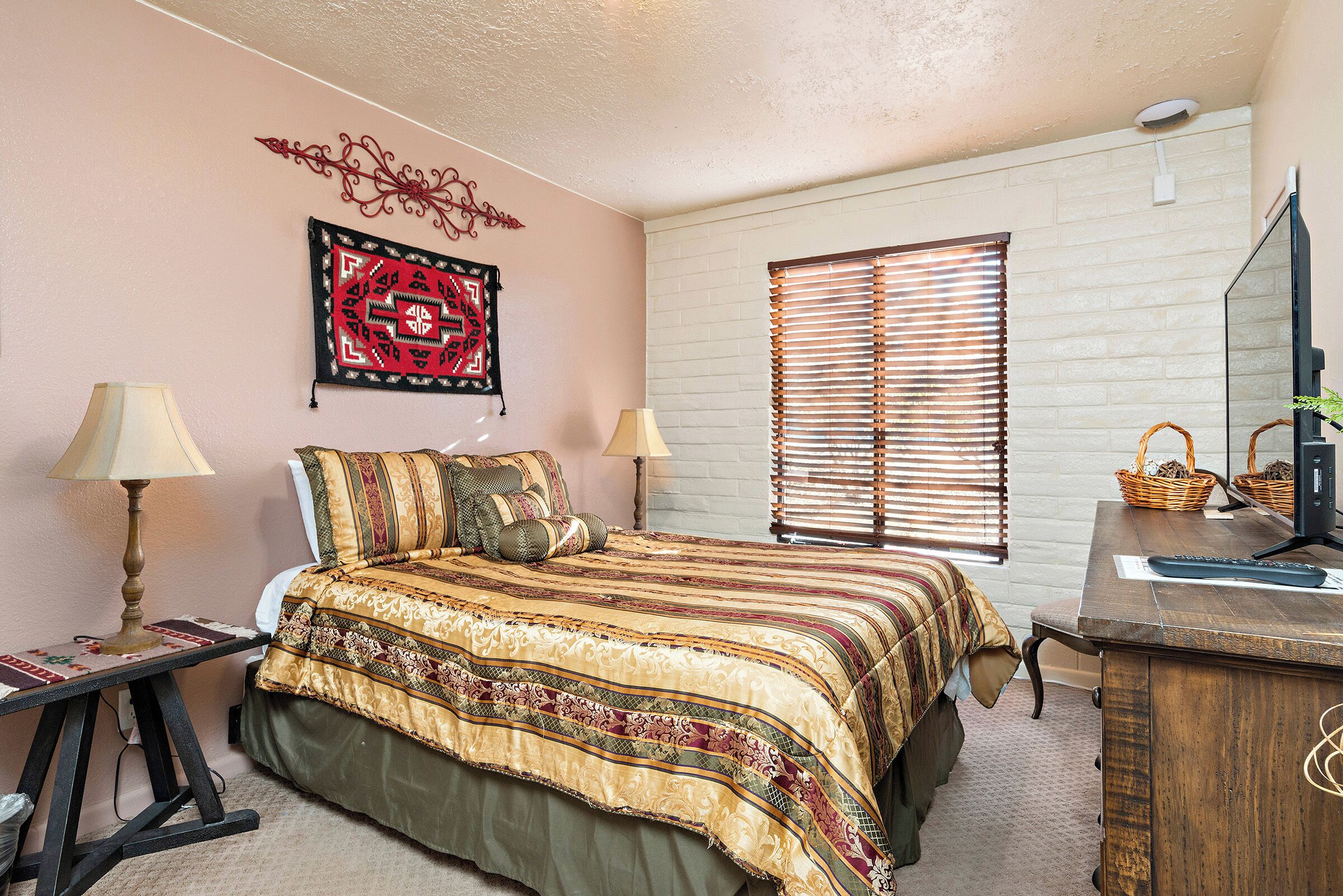 A bedroom with a bed, two bedside lamps, a dresser, a mirror, a window with blinds, and a wall decoration.