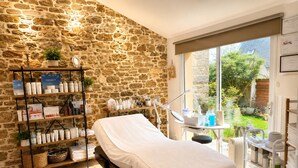 A room with a stone wall, a bed, a window with a view of greenery, and shelves with various items.