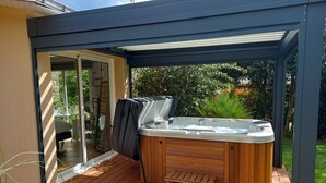 Un jacuzzi en un espacio al aire libre cubierto, con una terraza de madera, una puerta de cristal que da a una habitación y follaje de fondo.