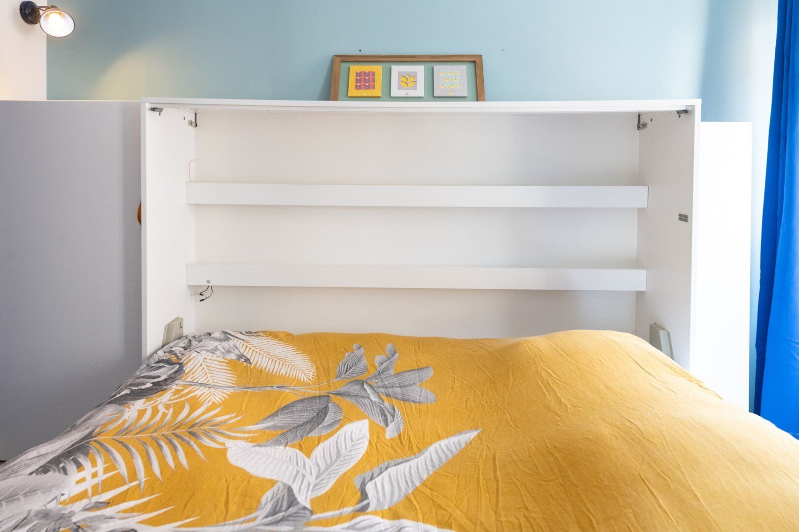 Een bed met een gele bloemenbedekking, een wit hoofdeinde en een plank erboven.