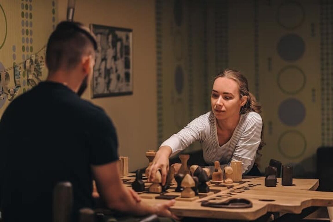 Une partie d’échecs est en cours sur une table en bois, avec un mur à motifs en arrière-plan.