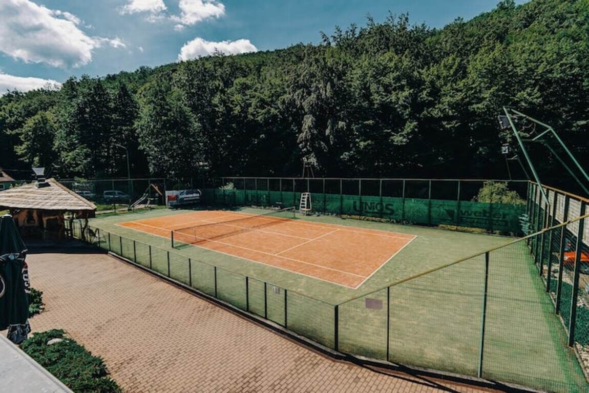 Un court de tennis avec un filet, entouré d’une clôture verte, et un chemin en briques menant à un bâtiment.