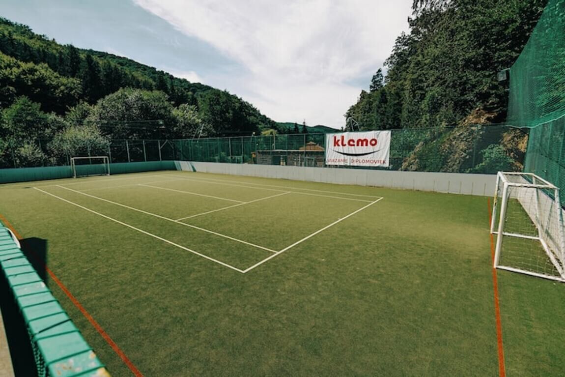 An outdoor tennis court with a net, surrounded by a green fence and trees.