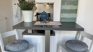 A modern kitchen with a dark countertop, white chairs with patterned cushions, a potted plant, and a blue teapot on a mat.