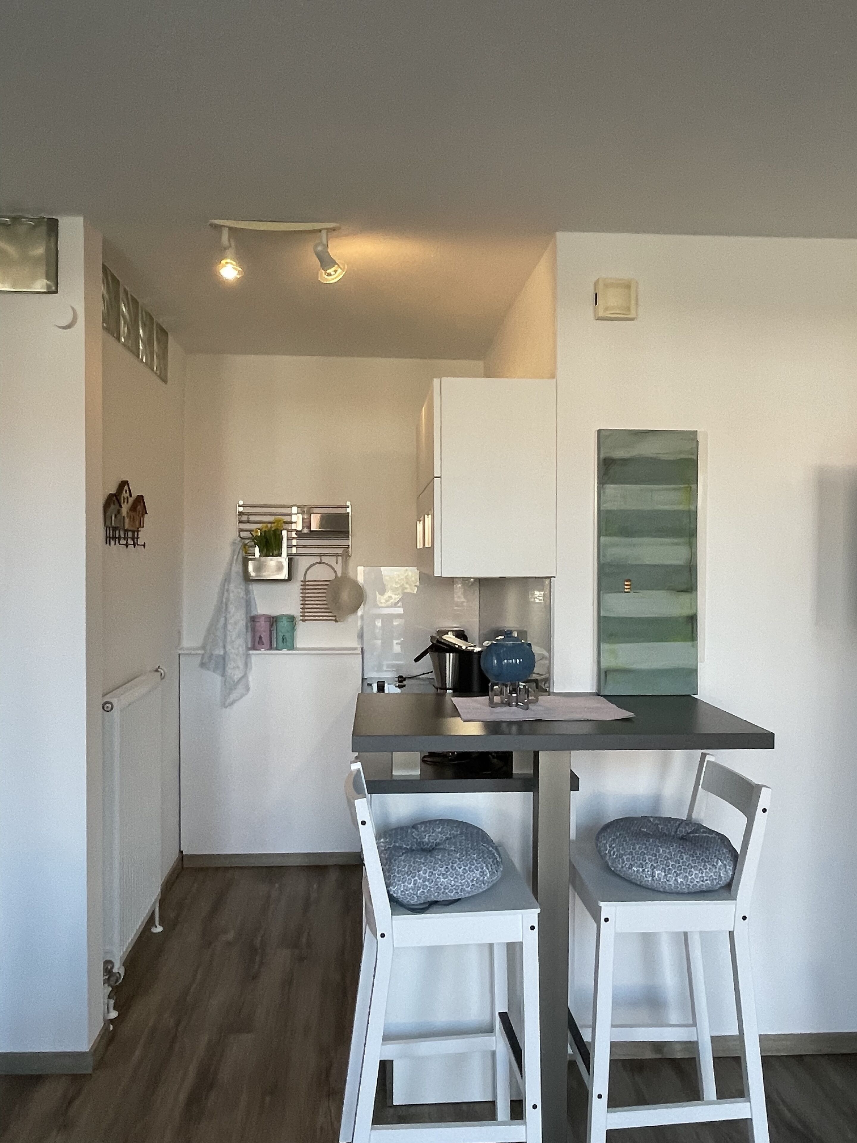 A compact kitchen with white cabinets, a small dining table, and two chairs. There is a refrigerator, a kettle, and a microwave on the counter.