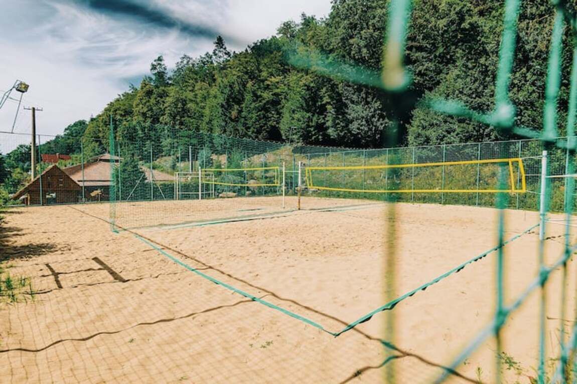 A sandy volleyball court enclosed by a net, surrounded by a fence and trees.