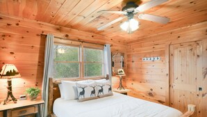 Un dormitori de cabana de fusta amb un llit, un ventilador de sostre, una finestra amb cortines, una taula de nit amb una làmpada i un penjador de paret.