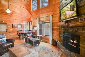 A cozy cabin interior with a fireplace, wooden walls, a dining area, and a large window. - Mountain view cabin | Hot tub | Peloton | Arcade | King | 11 mile to Blue Ridge (Mineral Bluff)