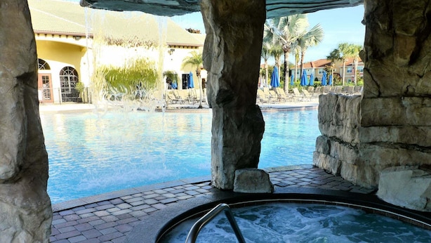 A hot tub area with a pool in the background, stone pillars, and a building with a yellow facade.