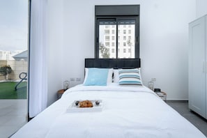 A modern bedroom with a large bed, a window with a view of buildings, and a tray with pastries on the bed.