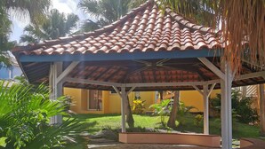 Um espaço ao ar livre coberto por um telhado de azulejos, rodeado por vegetação e um edifício ao fundo.