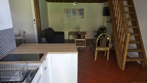 Cozinha compacta com pia, fogão e pequena geladeira. Há uma mesa de madeira e uma cadeira na área de refeições. Uma escada de madeira está encostada na parede.