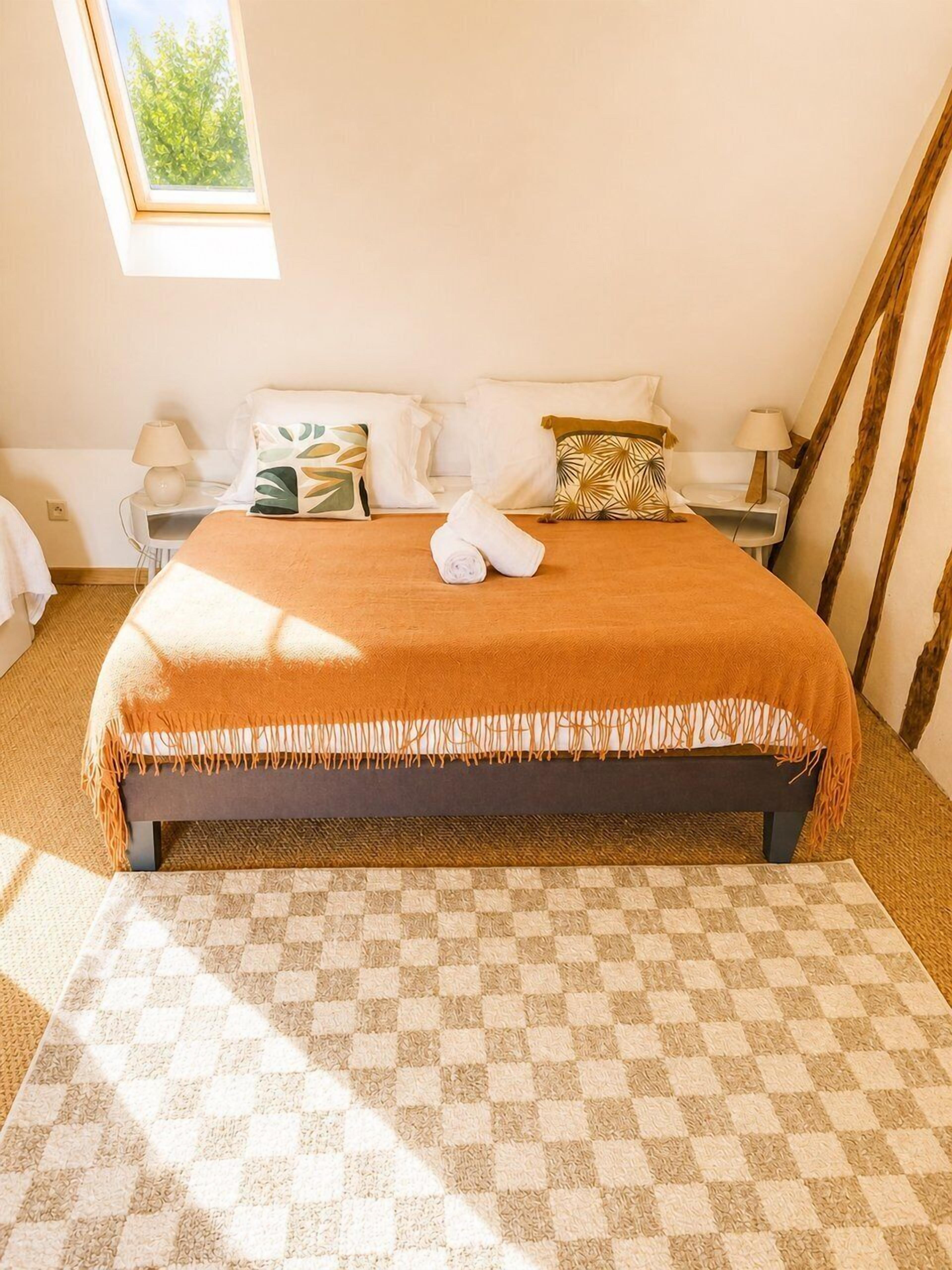 Une chambre avec un sol à carreaux, un lit recouvert d’une couverture marron et une fenêtre donnant sur un paysage verdoyant.