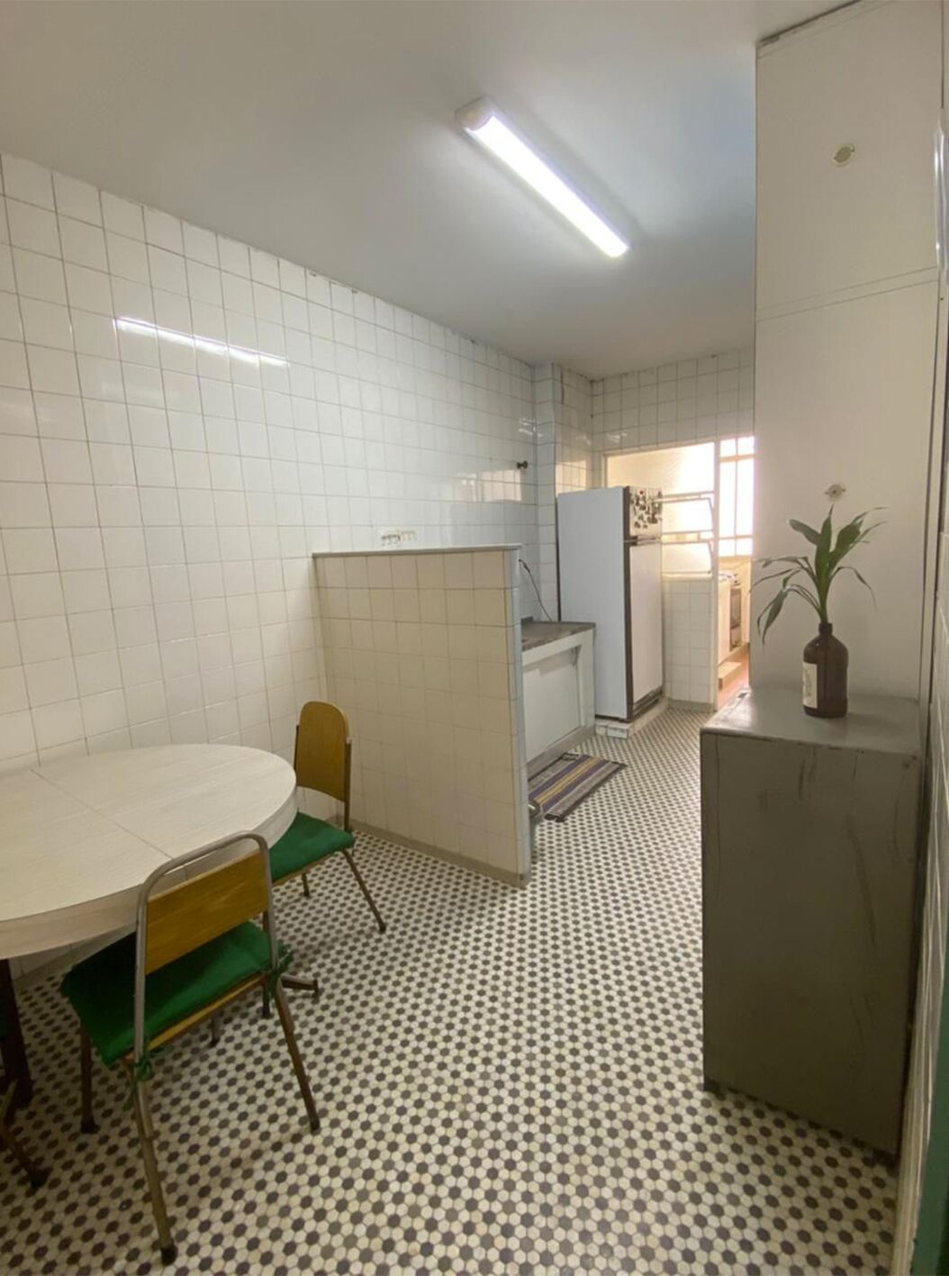 A small, tiled kitchen with a round table and chairs, a refrigerator, and a potted plant.