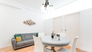 A dining area with a round table, white chairs, a gray sofa with green and yellow cushions, and a wall-mounted light fixture. - GuestReady Economy - Gold Leaf GF (Porto)