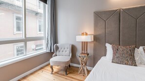 Ein Hotelzimmer mit großem Fenster, einem gepolsterten Sessel, einem Nachttisch mit Lampe, einem Bett mit gemusterten Kissen und einem Kopfteil mit geometrischem Muster.
