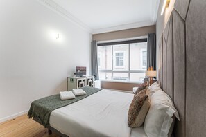 A hotel room with a bed, a desk with a computer, a window with curtains, and a lamp.