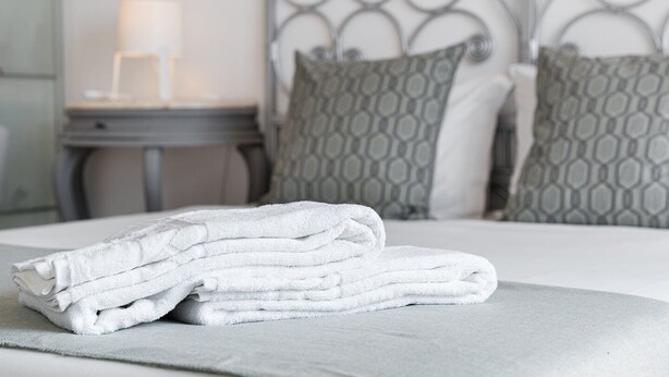 A neatly made bed with a stack of white towels, a grey patterned headboard, and a bedside table with a lamp. - GuestReady  Economy - Gold Leaf 2B (Porto)