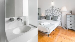 A modern bathroom with a large sink, mirror, and a bottle of soap. The room features a bed with a metal headboard, patterned pillows, and a bedside table with a lamp. - GuestReady  Economy - Gold Leaf 2B (Porto)