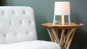Un fauteuil blanc à dossier capitonné et une table d’appoint en bois surmontée d’une lampe blanche.