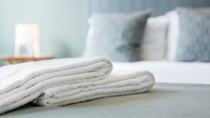 Une serviette blanche, soigneusement pliée, est posée sur un lit recouvert d’une couverture grise, dans une chambre dont le mur est également gris et qui comprend une table de chevet avec une lampe.