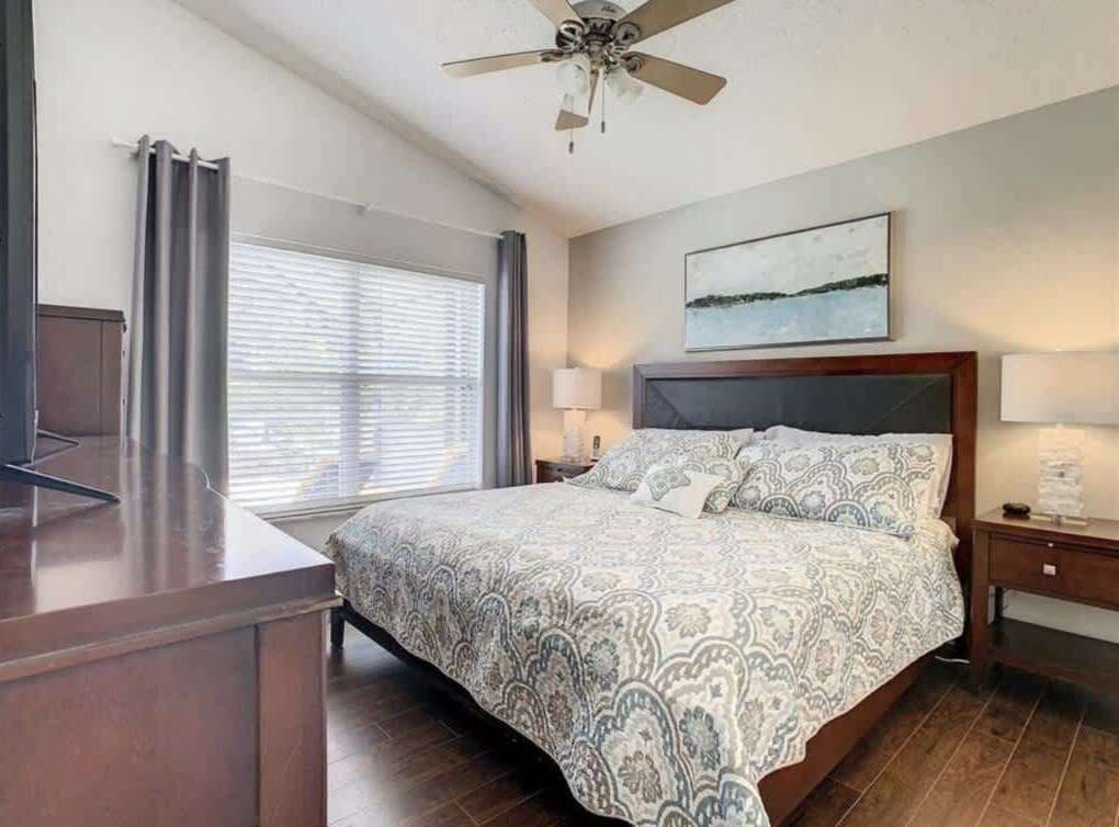 Ein Schlafzimmer mit Bett, Deckenventilator, Fenster mit Jalousien, Nachttisch und einem Gemälde an der Wand.