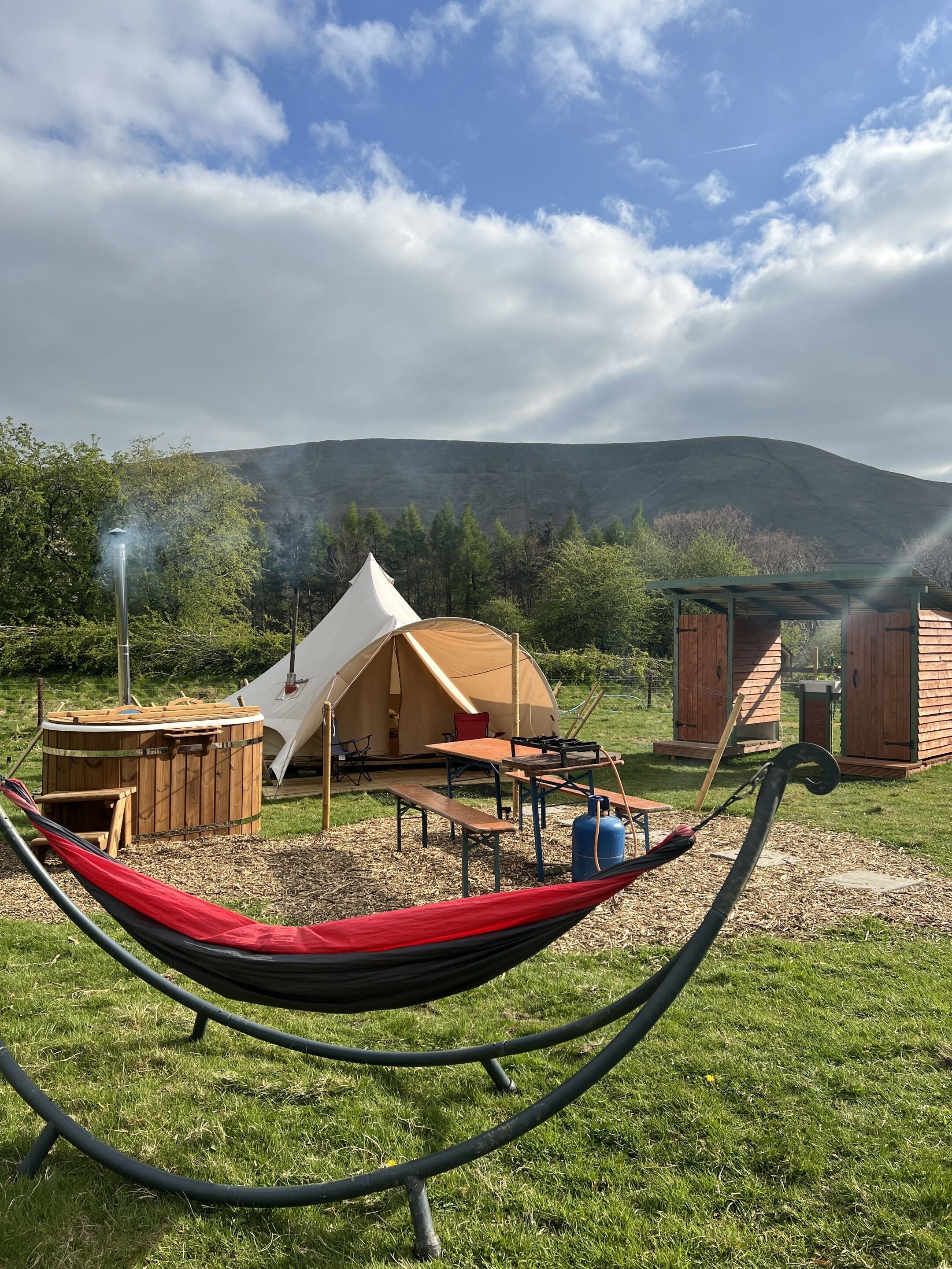 A campsite with a tent, a wooden hot tub, a hammock, and a barbecue area.