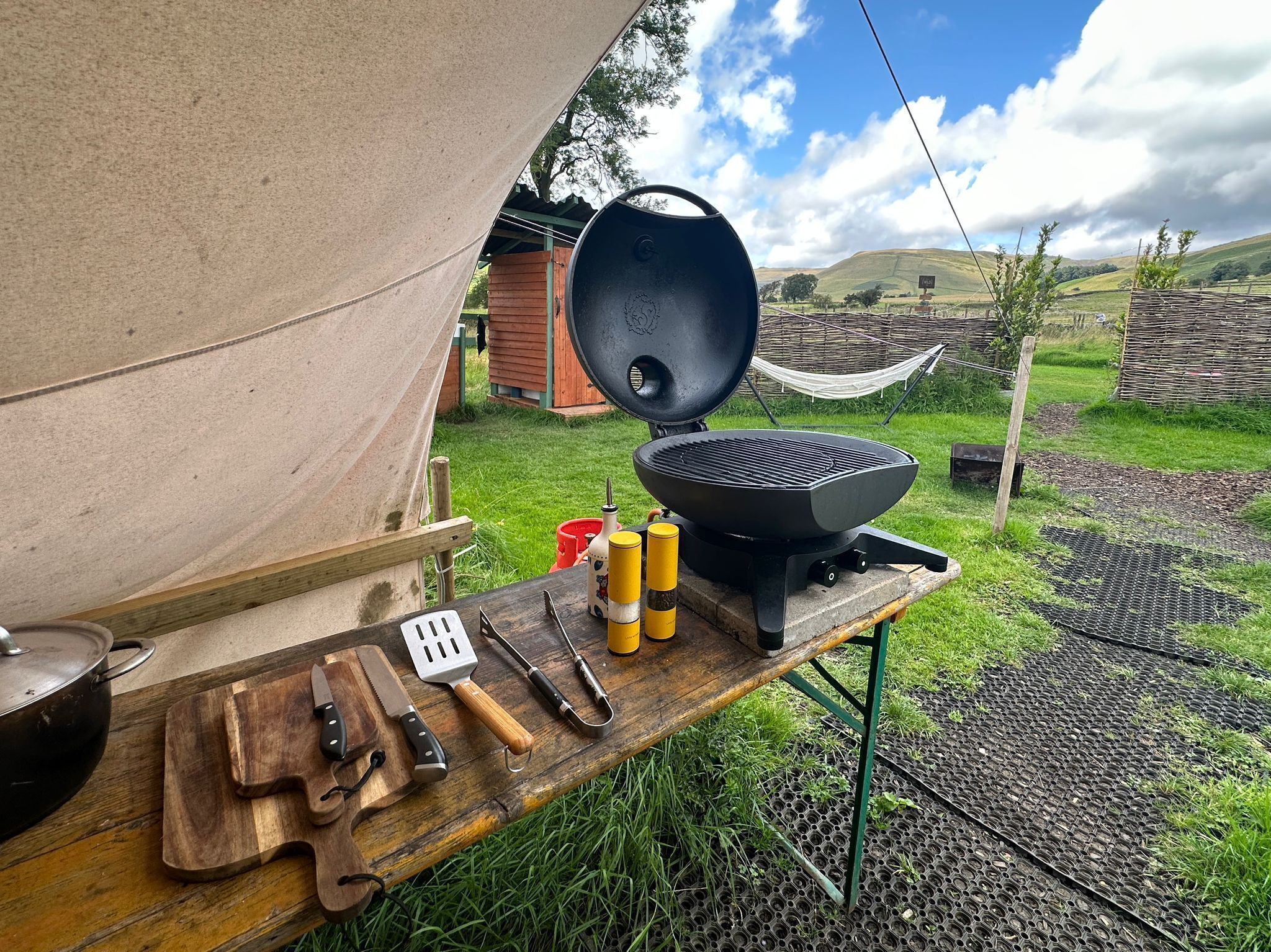 A portable grill set up outdoors with cooking utensils and a cutting board on a wooden table.