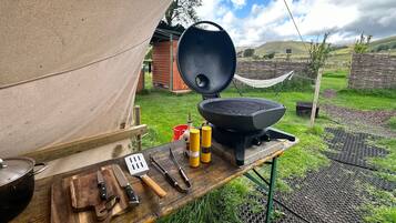 A portable grill set up outdoors with cooking utensils and a cutting board on a wooden table.