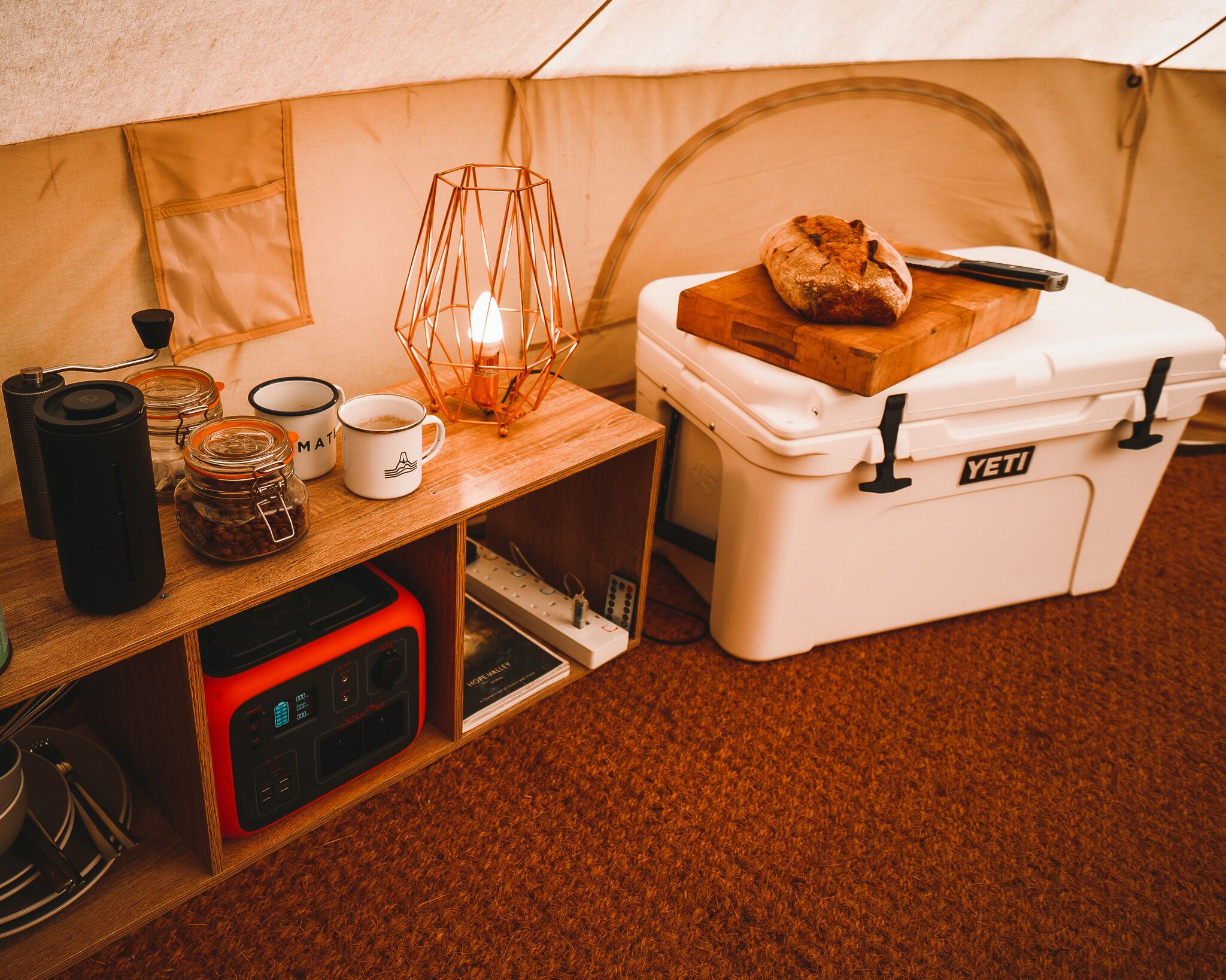 A tent interior with a table holding a coffee mug, a candle, and a loaf of bread. A portable stove and a white storage box labeled "YETI" are also visible.