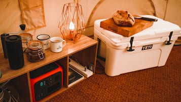 A tent interior with a table holding a coffee mug, a candle, and a loaf of bread. A portable stove and a white storage box labeled "YETI" are also visible.