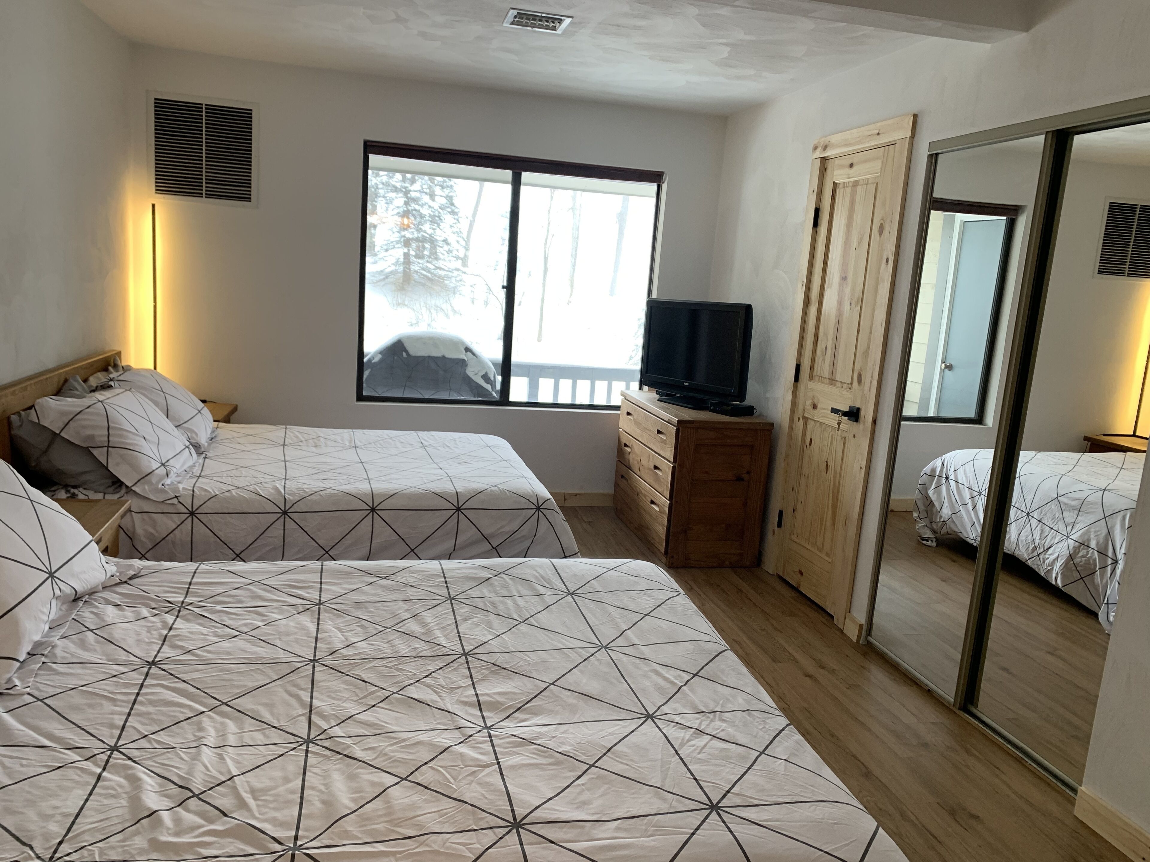 A bedroom with two beds, a TV on a wooden dresser, and a large window with a view of trees.
