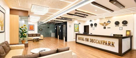 A hotel reception area with a reception desk, sofas, a coffee table, and a wall with clocks and awards.