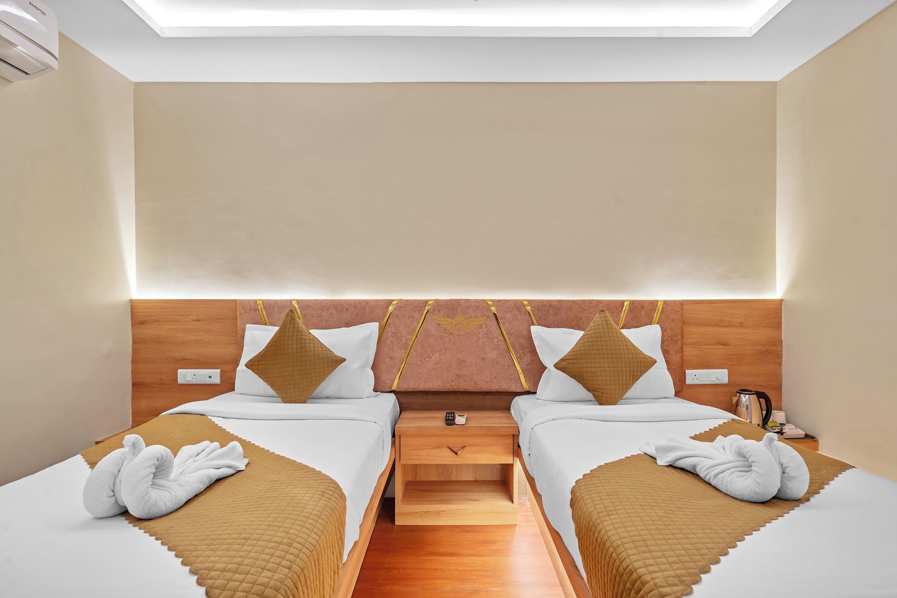 Habitación de hotel con dos camas, cabecera de madera, mesita de noche con tetera y aire acondicionado de pared.