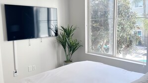 A bedroom with a large bed, a TV mounted on the wall, a window with blinds, and a potted plant. - Cute Poolside Unit, 2 Miles from the Beach (Marina del Rey)