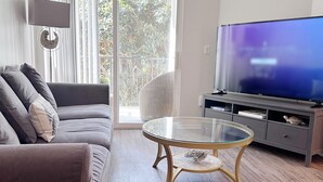 A modern living room with a sofa, a glass coffee table, a flat-screen TV, and a sliding glass door leading to a balcony. - Cute Poolside Unit, 2 Miles from the Beach (Marina del Rey)