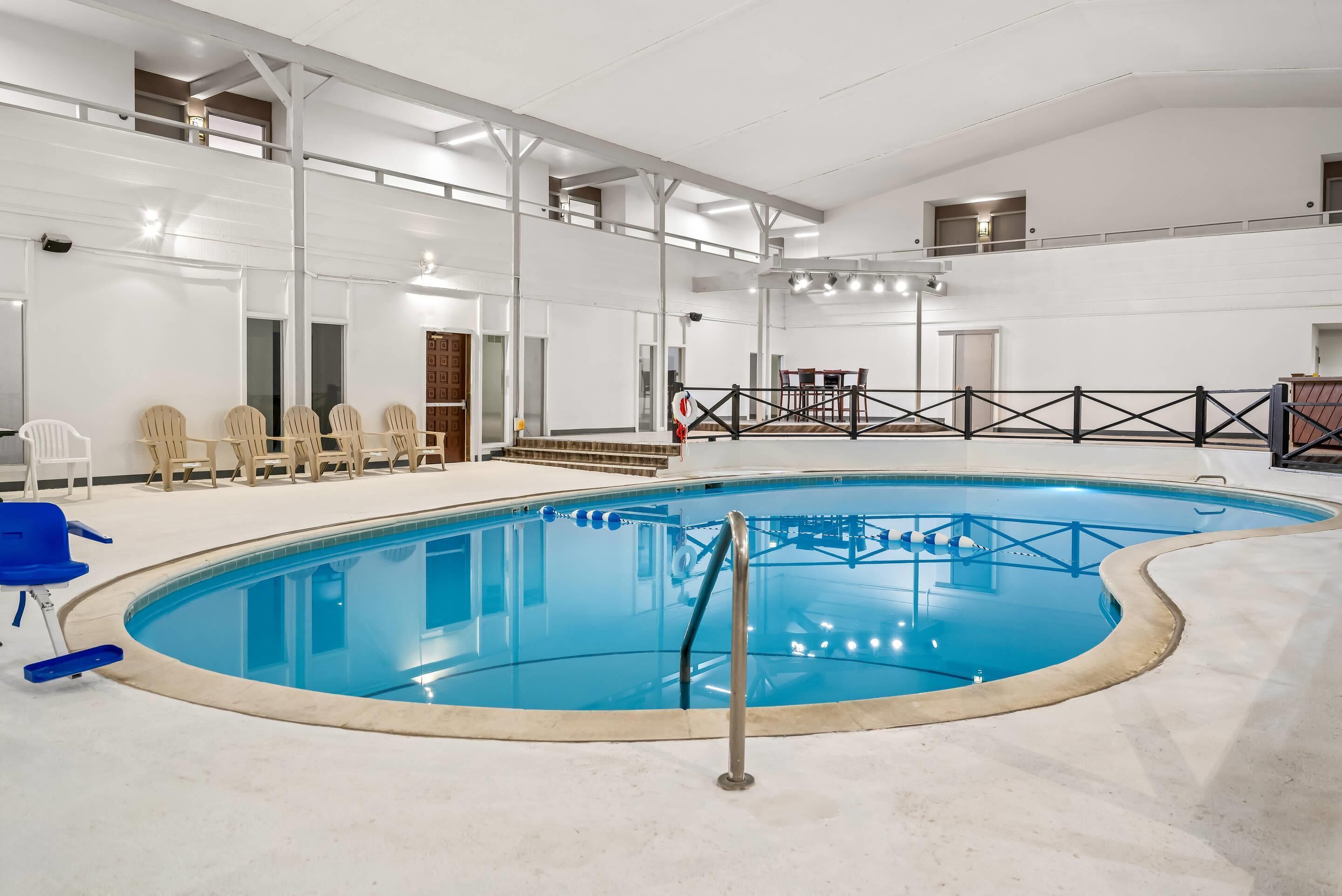 Une piscine intérieure avec un escalier en métal, un espace détente avec des chaises et un plafond haut avec des poutres apparentes.