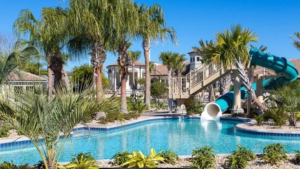Une piscine avec un toboggan aquatique, des palmiers et, au fond, un bâtiment avec un toit recouvert de tuiles.