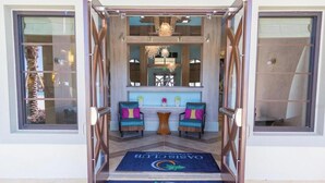 L’entrée d’un hôtel, avec un tapis bleu orné du logo du Oasis Club, deux fauteuils colorés, une petite table et un lustre.
