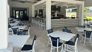 Un espace extérieur aménagé pour les repas, avec des tables et des chaises blanches, un bar doté d’un comptoir et des ventilateurs de plafond.