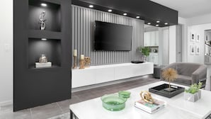 Un salon moderne avec un grand téléviseur à écran plat fixé au mur, lequel est recouvert de panneaux verticaux. On y trouve également un canapé gris, une table basse blanche agrémentée d’objets décoratifs et un fauteuil gris.