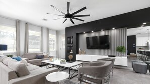 Un salon moderne avec un canapé, une table basse et des fauteuils. Un téléviseur est fixé au mur, et on aperçoit un coin cuisine au fond de la pièce.