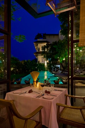 An outdoor dining area with a table set for two, overlooking a pool and a building with a balcony.