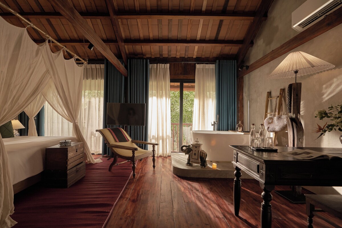 A bedroom with a wooden ceiling, a bed with a canopy, a chair, a table with a lamp, and a window with curtains.