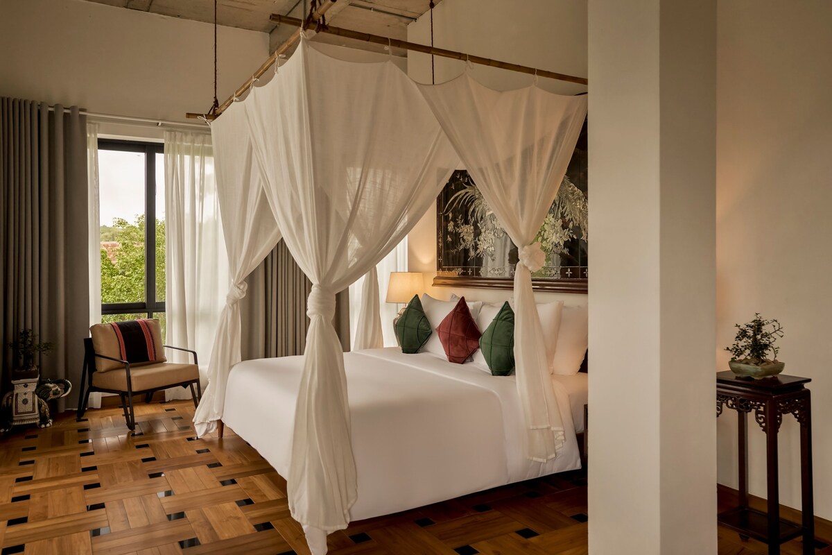 A four-poster canopy bed with a view of greenery outside, a bedside table with a lamp, and a chair with cushions.